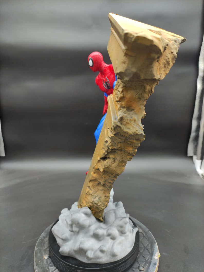 Pu&ograve; includere: Una action figure di Spider-Man rossa e blu &egrave; in posa mentre scala una formazione rocciosa rotta, grigia e marrone. La figura &egrave; in piedi su una base grigia e nera.