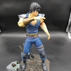 Puede incluir: Una figura de acción detallada de un personaje con cabello negro, una camisa azul y pantalones marrones. La figura está de pie sobre una base rocosa con una pose dramática.