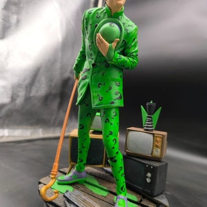 May include: A green and black action figure of The Riddler from Batman, holding a green top hat and a cane. The figure is standing on a wooden base with a television set and other props.