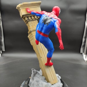 Pu&ograve; includere: Una action figure di Spider-Man che scala una colonna di pietra. La figura &egrave; in posa con una gamba sulla colonna e un braccio teso. La figura &egrave; posizionata su una base con un effetto nuvola grigia.