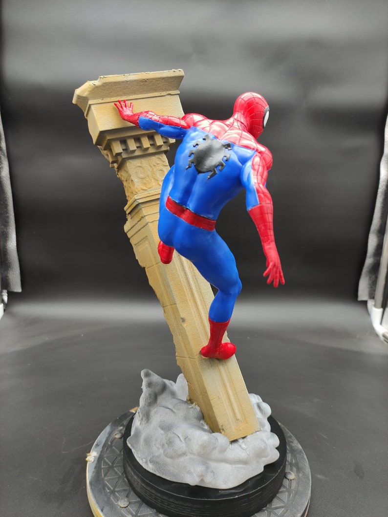 Pu&ograve; includere: Una action figure di Spider-Man che scala una colonna di pietra. La figura &egrave; in posa con una gamba sulla colonna e un braccio teso. La figura &egrave; posizionata su una base con un effetto nuvola grigia.