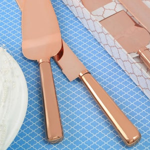 Puede incluir: Un juego de dos servidores de pastel de oro rosa. Los servidores están hechos de metal y tienen un acabado liso y pulido. Los servidores están en una caja blanca con un patrón decorativo.