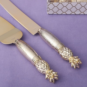 Könnte beinhalten: Goldenes Ananas-Kuchenmesser-Set. Das Set enthält ein gezahntes Messer und einen Streicher mit goldenen Ananasgriffen.