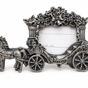 May include: A silver-toned metal place card holder shaped like a horse-drawn carriage. The frame features ornate floral detailing, columns, and wheels. A white insert is included with the printed text M. and Table No. with blank lines for writing.
