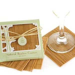 Peut inclure: Un ensemble de quatre dessous de verre en bambou naturel, présenté dans une boîte vert clair attachée avec un ruban et une petite étiquette indiquant "For You". Un verre à vin est posé sur l'un des dessous de verre. Les dessous de verre sont faits de fines lattes de bambou.