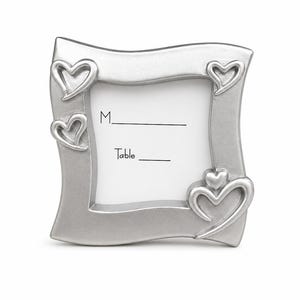 May include: A silver place card holder with a wavy frame and heart-shaped embellishments. The holder has space for a name and table number, with the words "M_" and "Table_" printed on the white insert.