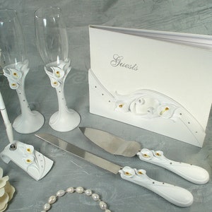 Może przedstawiać: Biały zestaw do serwowania tortu weselnego z białym notesem dla gości, dwoma białymi kieliszkami do szampana z białymi wzorami lilii i białym stojakiem na długopis z białym wzorem lilii. Notes dla gości ma napis "Guests" na okładce.