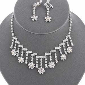 May include: A silver-coloured necklace and earring set. The necklace features a series of vertical, dangling crystal accents, each with a flower-shaped cluster of crystals. The matching earrings have a similar design, with a flower-shaped crystal at the bottom.