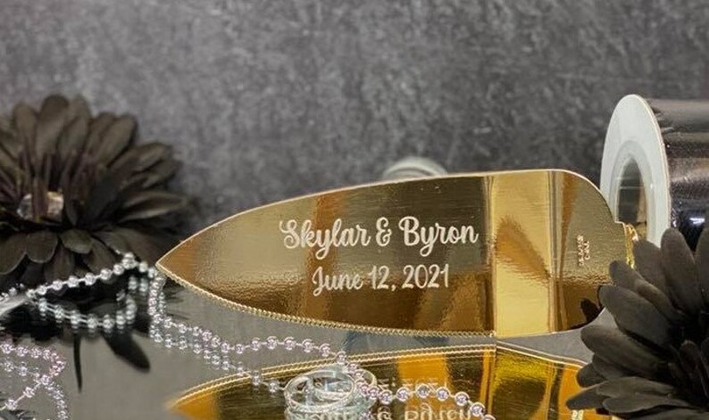 K&ouml;nnte beinhalten: Ein goldfarbener Kuchenspachtel mit der Aufschrift "Skylar & Byron June 12, 2021".