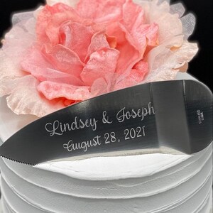 May include: A silver cake knife with the inscription "Lindsey & Joseph August 28, 2021" on a white cake with a pink flower on top.