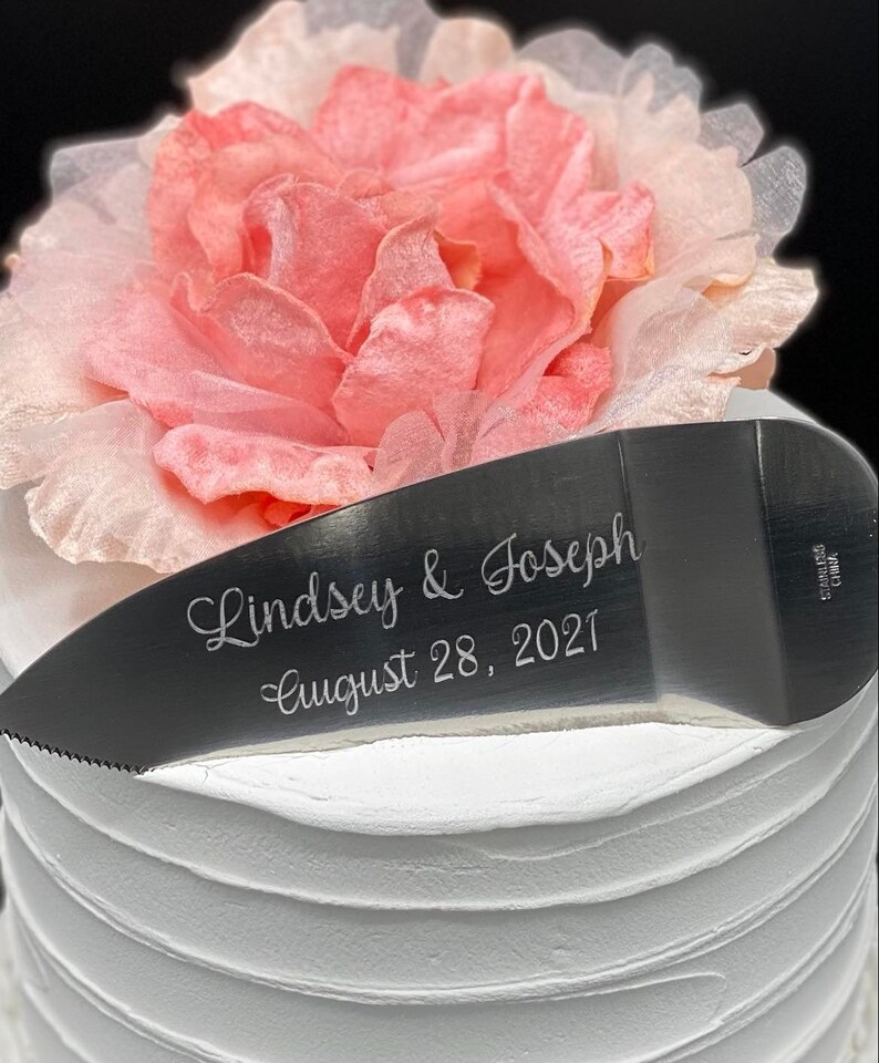 May include: A silver cake knife with the inscription "Lindsey & Joseph August 28, 2021" on a white cake with a pink flower on top.