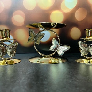 Puede incluir: Tres candelabros dorados con diseños de mariposas. Cada candelero tiene una base redonda y una copa para una vela. El candelero del centro es más alto y tiene dos mariposas.