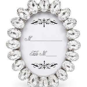 May include: An oval-shaped table number card holder featuring a silver-toned frame encrusted with clear, faceted oval crystals. The white insert card displays black printed text reading M and Table No. with decorative flourishes at the top and bottom.