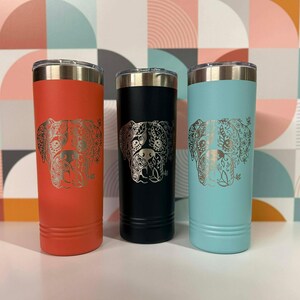 May include: Three insulated tumblers in coral, black, and teal. Each tumbler features a detailed silver dog face design. The tumblers have a stainless steel rim and lid. The background has a colorful geometric pattern.