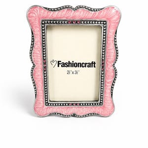 May include: A pink and silver photo frame with a decorative, scalloped edge. The frame has a pink enamel finish with silver accents and is embellished with small crystals. The frame has the text "Fashioncraft" and the dimensions 6.35cm x 8.89cm.
