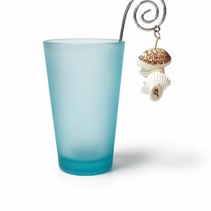 Könnte beinhalten: Ein mattiertes, hellblaues Glas mit einer dekorativen Metallspirale und einem Muschelanhänger. Das Glas hat eine einfache, zylindrische Form. Der Anhänger zeigt zwei Muscheln, eine davon mit braunem und weißem Muster.