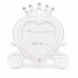 May include: A white place card holder in the shape of a heart, topped with a crown. It has space for a name and table number, adorned with roses and crystals. The design evokes a fairytale carriage, perfect for weddings.