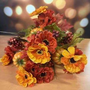 May include: A fall-themed floral arrangement featuring orange and yellow flowers with black centers, small pumpkins, and red berries. The arrangement includes green leaves and is set against a blurred background.