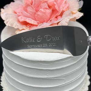 May include: A silver cake knife with a white handle. The knife is engraved with the text "Katie & Drew September 25, 2021".