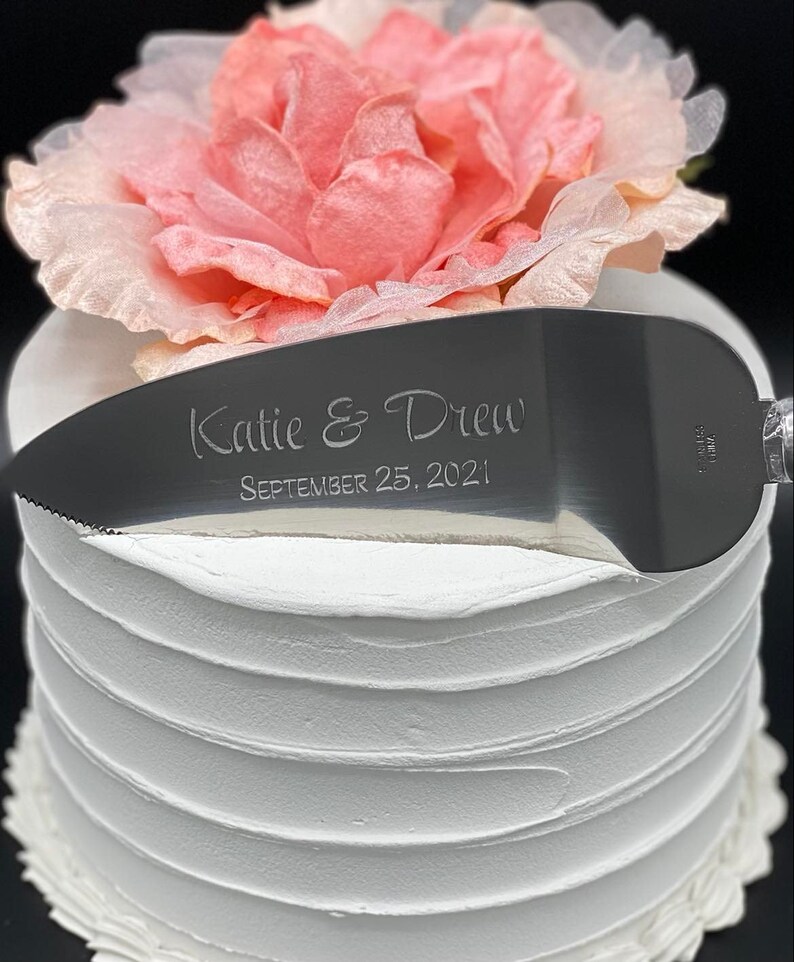 May include: A silver cake knife with a white handle. The knife is engraved with the text "Katie & Drew September 25, 2021".