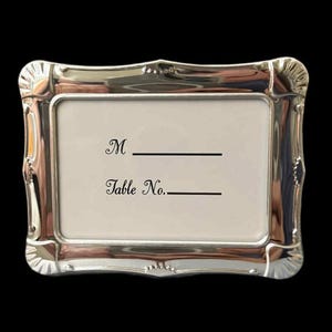 May include: A silver-colored rectangular picture frame with a decorative, ornate border. The frame holds a white card with the words "M" and "Table No." printed in black. The frame is set against a black background.