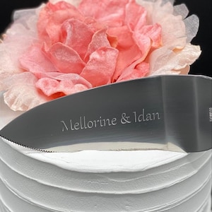 May include: A silver cake knife with a serrated edge and the inscription "Mellorine & Idan" on the blade. The knife is resting on top of a white cake decorated with a pink flower.