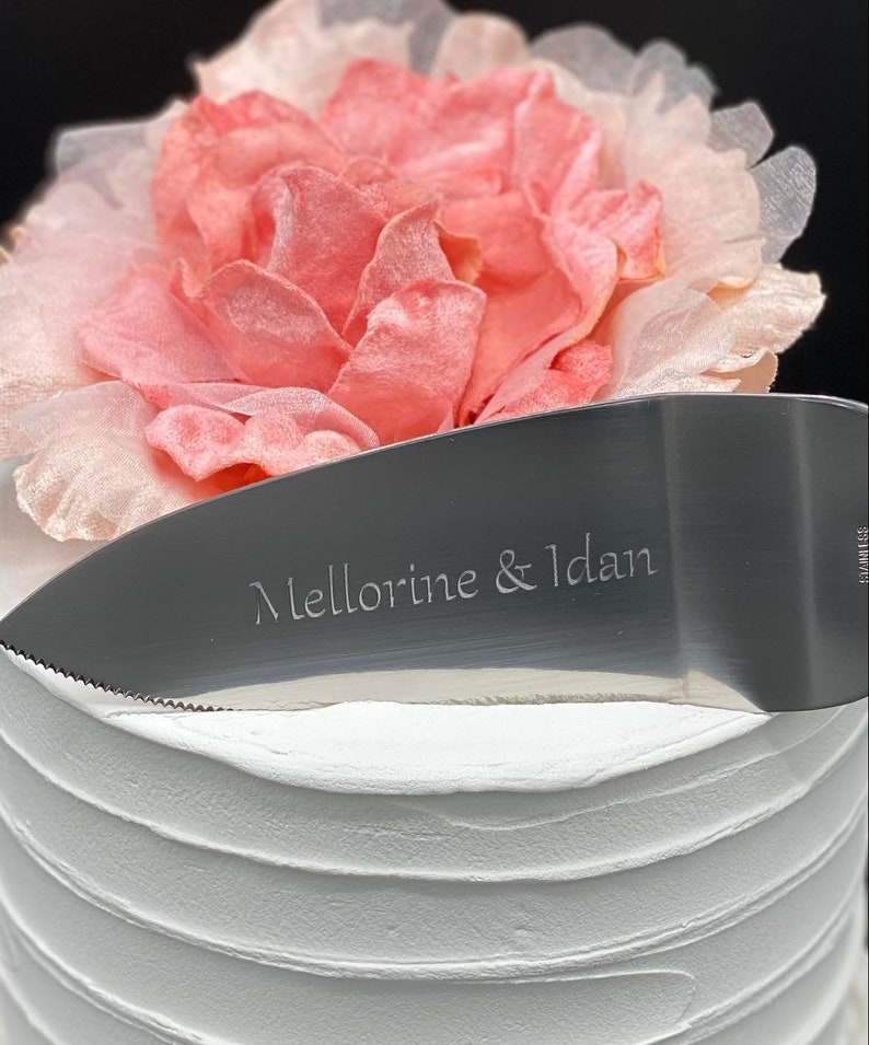 May include: A silver cake knife with a serrated edge and the inscription "Mellorine & Idan" on the blade. The knife is resting on top of a white cake decorated with a pink flower.