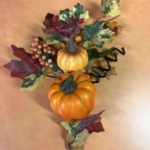 May include: An arrangement of two orange pumpkins with green stems, surrounded by colorful fall leaves and berries. The leaves are in shades of red, green, and yellow, creating a festive autumn display.