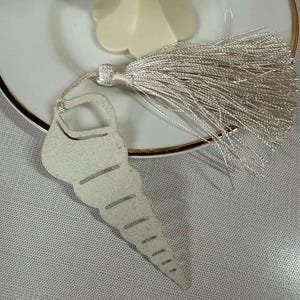 May include: A seashell-shaped bookmark with a silver tassel. The bookmark is a light, neutral color and has a cut-out design. The tassel is made of many strands of silver thread. The bookmark rests on a white plate with a gold rim.