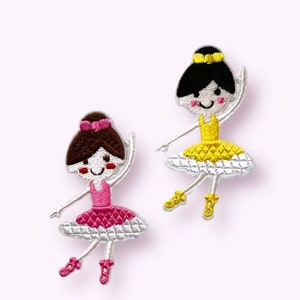 May include: Two embroidered patches of cartoon ballerinas. One ballerina wears a pink dress and a brown bun with a pink bow. The other ballerina wears a yellow dress and a black bun with a yellow headband. Both have white tutus and are smiling.