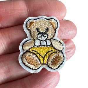 May include: A small embroidered iron-on patch featuring a cartoon teddy bear wearing yellow shorts. The bear is sitting with its paws in front of it.
