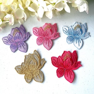 May include: Five embroidered lotus flower patches in purple, pink, blue, gold, and red. Each patch features detailed stitching and a small cluster of gold beads in the centre. White and yellow flowers are in the background.