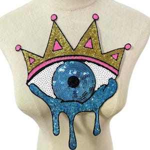 May include: A sequined patch featuring a stylized eye design with a crown. The crown is gold with pink accents, and the eye is blue with white and black details. The design includes dripping blue sequins below the eye.