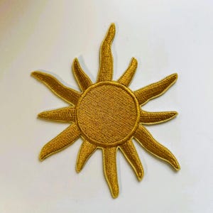 Könnte beinhalten: Gestickter Aufnäher mit einem goldenen Sonnendesign. Die Sonne hat eine kreisförmige Mitte mit ausstrahlenden Strahlen. Der Aufnäher ist aus goldenem Faden gefertigt und dient wahrscheinlich dekorativen Zwecken.