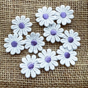 May include: White daisy flower patches with purple centres. These embroidered appliques feature detailed petal stitching. Ideal for adding a floral touch to clothing, accessories, or craft projects. The patches are arranged on a textured, brown surface.