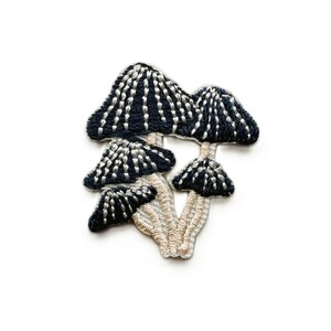 May include: Embroidered patch depicting a group of stylized mushrooms. The mushroom caps are navy blue with white detailing, and the stems are a light beige colour. The patch is designed to be sewn onto fabric.