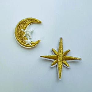 May include: Two embroidered patches featuring celestial designs. One patch is a gold crescent moon with two small white stars attached to its inner curve. The second patch is a four-pointed star shape in a metallic gold thread with white borders.