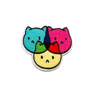 May include: An embroidered patch featuring three stylised cat heads arranged in a cluster. The top left cat is bright blue, the top right is hot pink, and the bottom centre cat is yellow. Each cat has simple black eyes and a small frown.