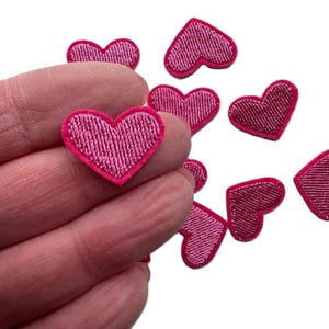May include: A collection of pink felt heart-shaped patches. The patches are approximately 1 inch in size and have a pink satin stitch around the edges.