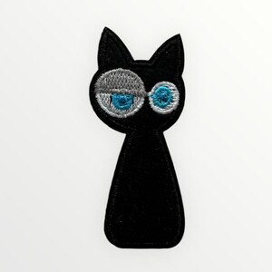 Könnte beinhalten: Schwarzer gestickter Bügelaufkleber mit einem Cartoon-Katzenmotiv mit großen blauen Augen und silbernem Umriss.