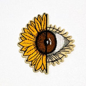 May include: An embroidered patch featuring a split design. The left side displays a vibrant yellow sunflower petal arrangement, while the right side depicts a detailed human eye with a brown iris and white sclera, framed by dark eyelashes.
