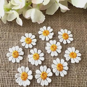Könnte beinhalten: Eine Ansammlung weißer Gänseblümchen-Blumenaufnäher mit gelben Zentren, angeordnet auf einer strukturierten, beigefarbenen Stoffoberfläche. Die gestickten Blumen sind in verschiedenen Größen und sind von weißen Blumen umgeben.