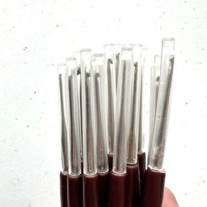 K&ouml;nnte beinhalten: Ein Satz durchsichtiger Plastik-H&auml;kelnadeln mit braunen Griffen. Die Nadeln sind geb&uuml;ndelt und einsatzbereit.