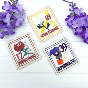 May include: Three embroidered iron-on patches with a white background and scalloped edges. The patches feature a white flower with a gold key, a red flower with the Eiffel Tower, and a blue bird with a red envelope. The patches have French phrases on them: "Bonne Courage", "Bonne Chance", and "Je Pense a Toi".