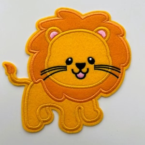 May include: A cheerful, embroidered lion patch in shades of yellow and orange. The lion has a smiling face with black whiskers and a pink mouth. The patch is likely for clothing or accessories.