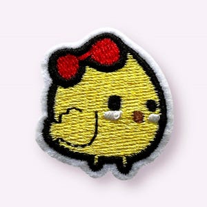 May include: An embroidered yellow chick with a red bow, black eyes, and a brown beak. The chick is standing on two small feet.