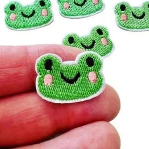 May include: Close-up of several green frog patches with black eyes and pink cheeks. The patches are embroidered with a white border and a smiling mouth. The patches are small and likely for clothing or craft projects.