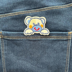 May include: A blue denim pocket with a light blue, yellow, and red embroidered bear patch. The bear has a smiling face and is peeking out of the pocket.