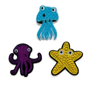 May include: Three embroidered patches featuring sea creatures: a blue jellyfish, a purple octopus, and a yellow starfish. Each patch has a black border and detailed stitching. The jellyfish has a textured cap and flowing tentacles.