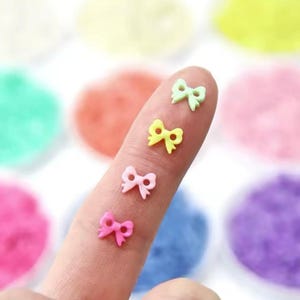 May include: Five pastel-colored plastic bows, each with a different color: pink, yellow, light blue, light green, and a darker shade of pink. The bows are arranged on a finger, with the smallest bow at the bottom and the largest bow at the top.